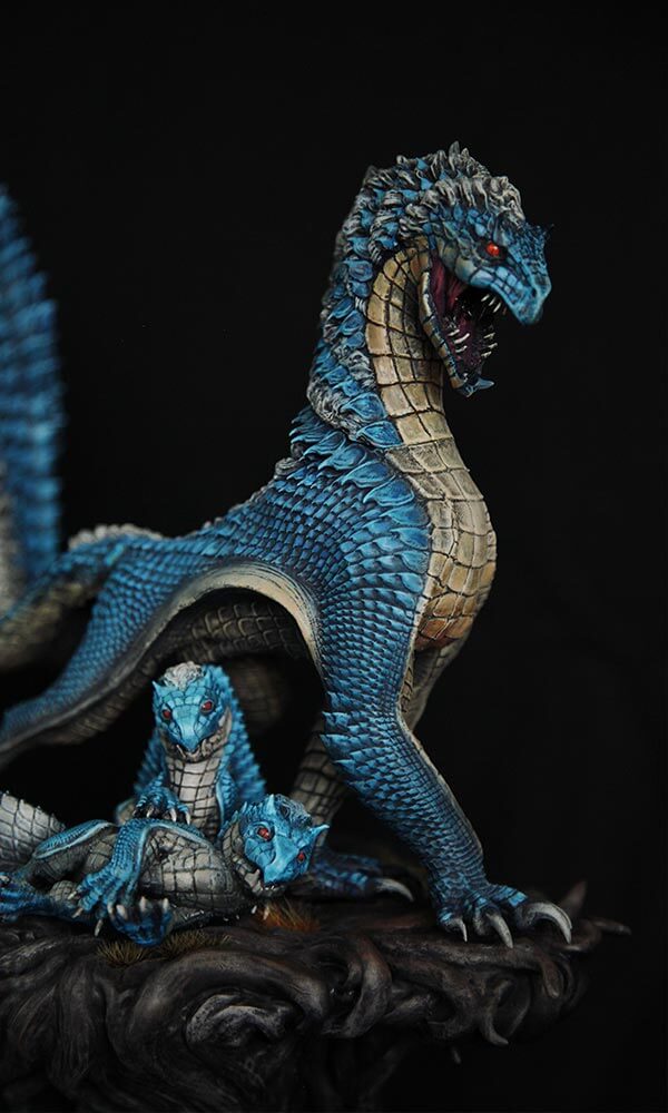Skulptur „Tobi Kadachi – Matriarch“ von Winton Afrić: blau schuppige Drachen-Kreatur mit geöffnetem Maul beschützt drei Jungtiere auf einem dunklen, wurzelartigen Sockel vor schwarzem Hintergrund.