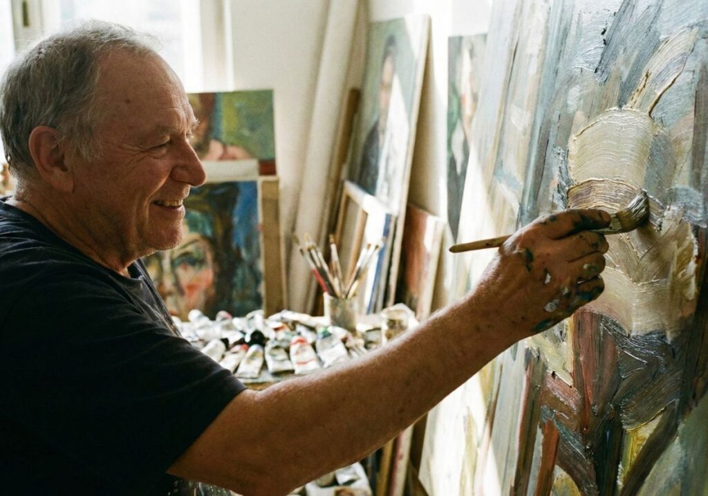 A close-up of an older artist with paint-smeared hands in a sunlit studio, smiling with visible confidence as he applies a bold, textured brushstroke to an oil painting.